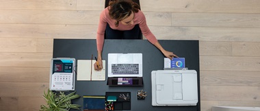 Blick von Oben auf einen schreibtisch mit Laptop und Drucker ein Den einer Frau sitzt.