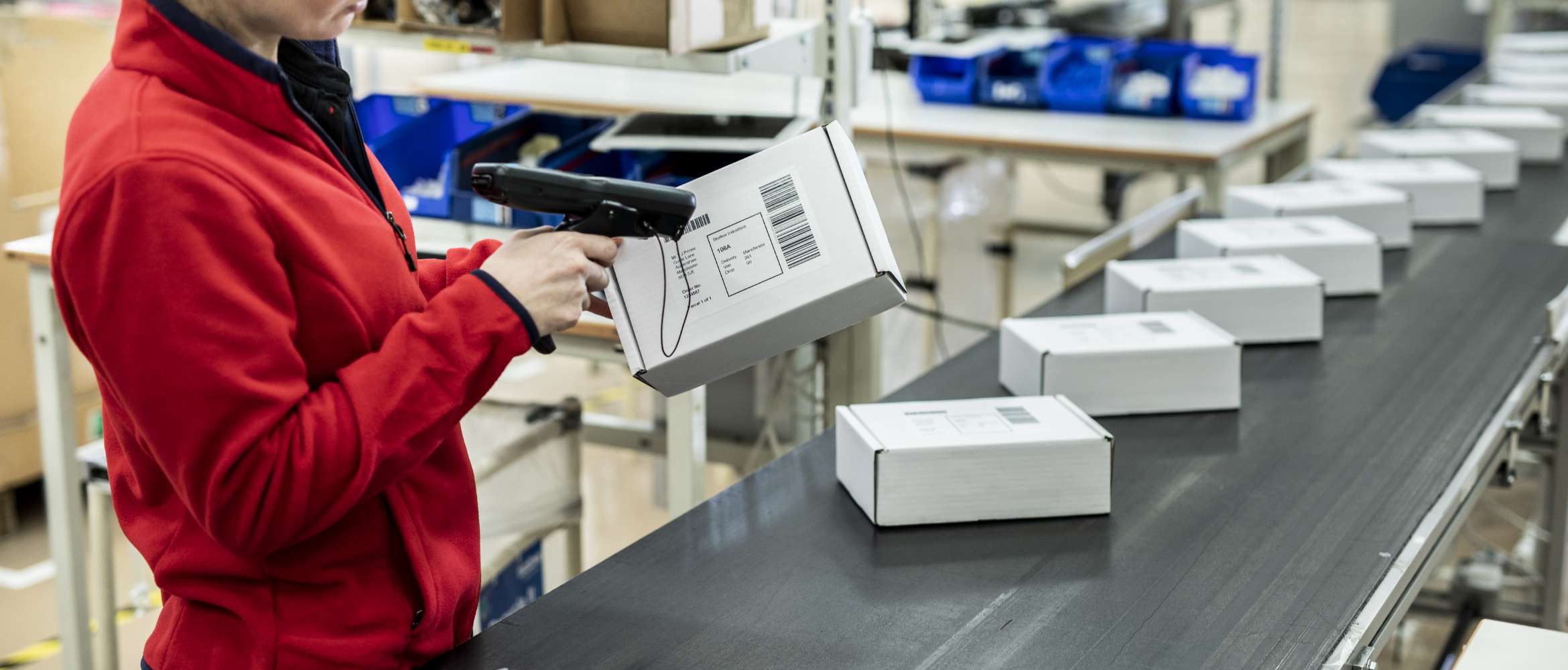 Ein Fließband in einer Fabrik wird mit verpackten Paketen gefüllt, während ein Arbeiter in dieser Transport- und Logistiklagerszene ein gedrucktes Barcode-Adressetikett scannt.