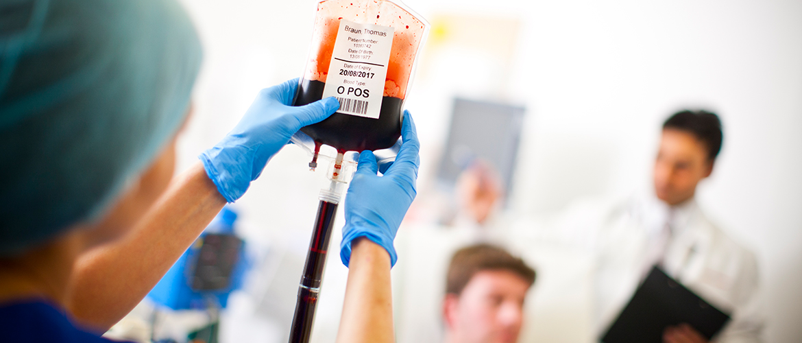 KRANGEPFLEGERIN KLEBT Barcode-Etikett auf Blutbeutel. Patient mit Arzt im Hintergrund.