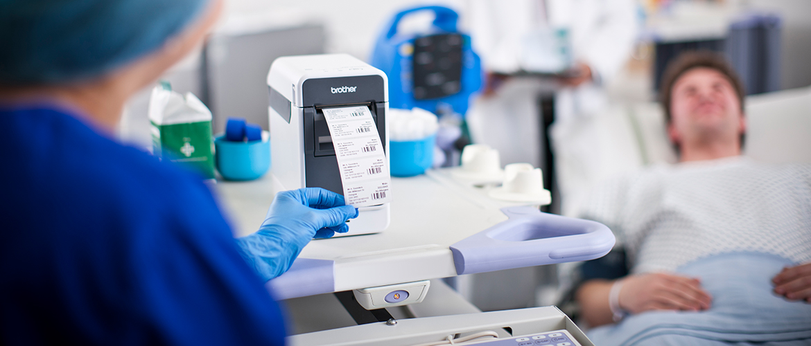 Krankenpfegerin Entnimmt Barcode-Etikett Aus Drucker mit Patienten im Hintergrund.