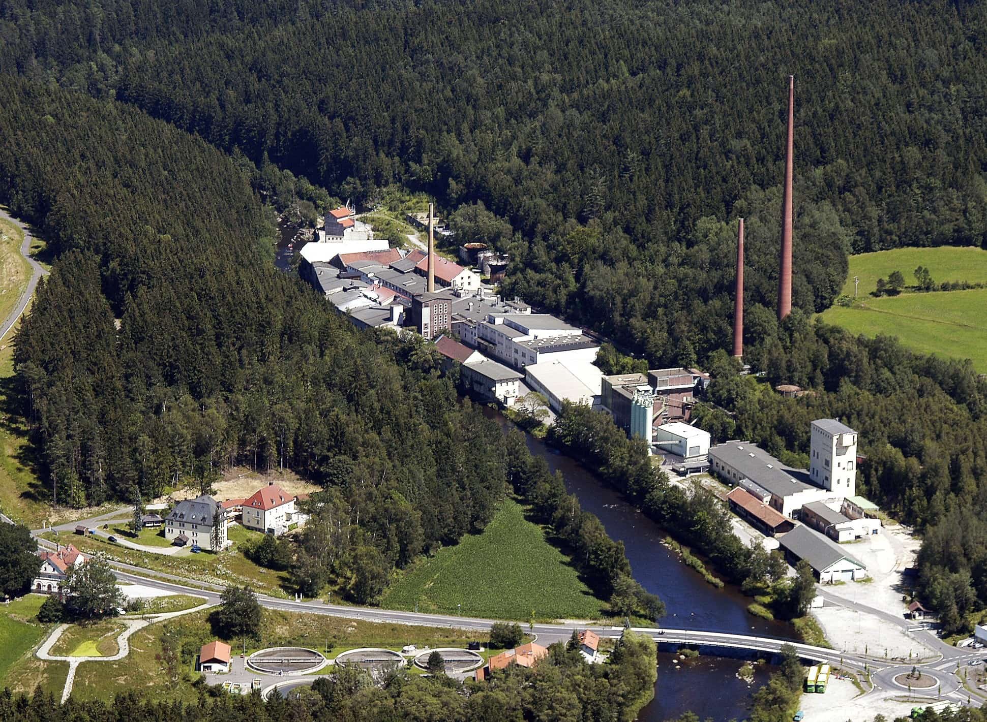 Fabrikgebäude an einem Fluss, welcher von einem Wald umgeben ist.