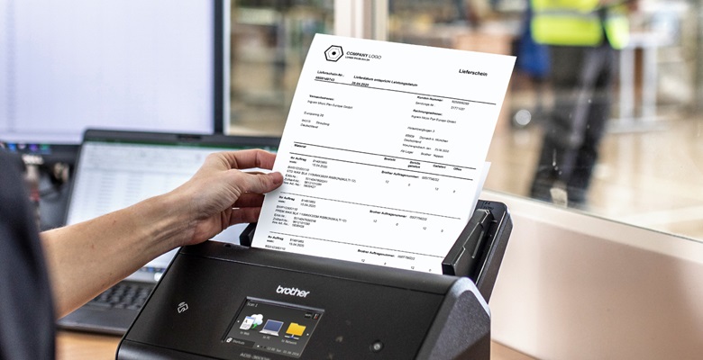 Frau im Schwarzen Polosht in Lagerbüro KommissionierListe mit Hand in DokumenteScanner ein. Laptop, Bildschirm und Lagerarbeit im Hintergrund