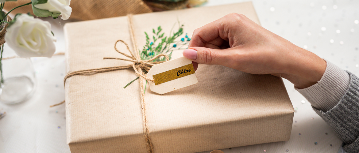 Frauenhand hält ein Etikett mit dem Namen Chloe darauf, das an einem Geschenk mit Band und braunem Papier befestigt ist