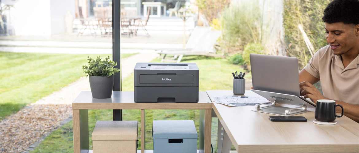 man sat at desk looking at laptop next to a printer and boxes on top of a sidetable with a garden in the background