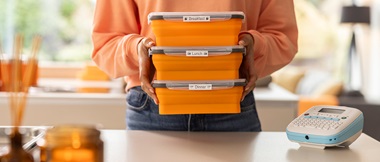 Eine Person hält in ihren Händen drei orangefarbene Lebensmittelboxen. Vor ihr auf dem Tisch liegt ein Btag Etikettendrucker. 