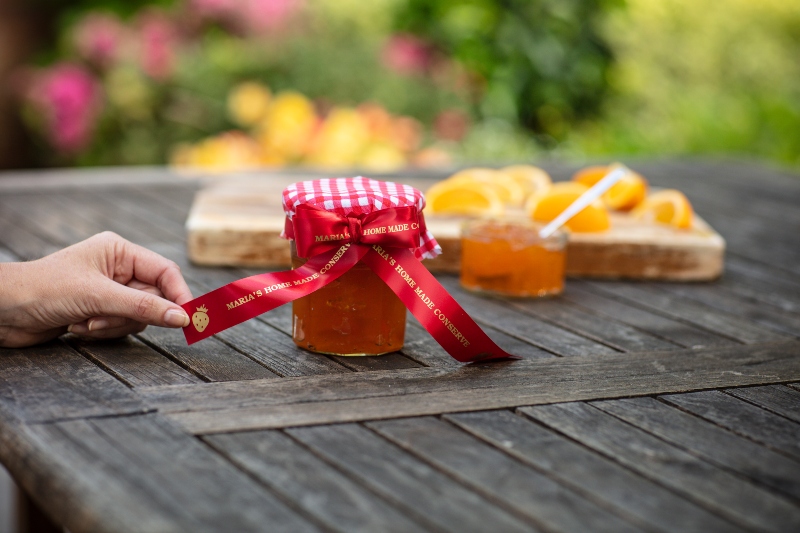 Hand, die ein rot bedrucktes Band hält, das an ein Marmeladenglas gebunden ist, auf einem Tisch mit geschnittenen Orangen im Hintergrund
