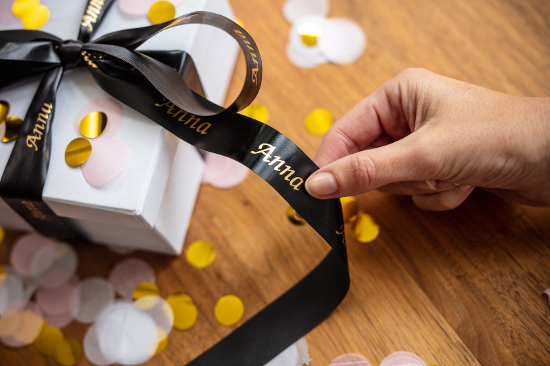 Eine Hand hält ein schwarzes und goldenes personalisiertes Band, das an eine weiße Schachtel gebunden ist, die mit gelbem und weißem Konfetti bedeckt ist.