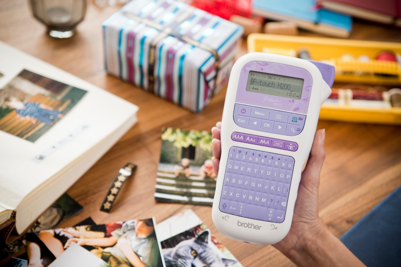 Eine Hand hält ein lilafarbenes P-touch-Etikettiergerät, im Hintergrund ein Tisch mit einer als Geschenk verpackten Schachtel, die mit einem bedruckten Band verschnürt ist, ein offenes Fotoalbum und einige Fotos