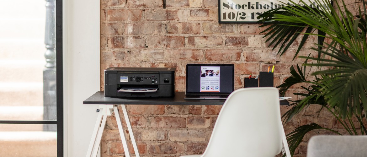 Ein Schreibtisch mit einem Drucker, einem Laptop und einigen Schreibwaren vor einer freiliegenden Backsteinwand und einer großen Grünpflanze auf der rechten Seite.