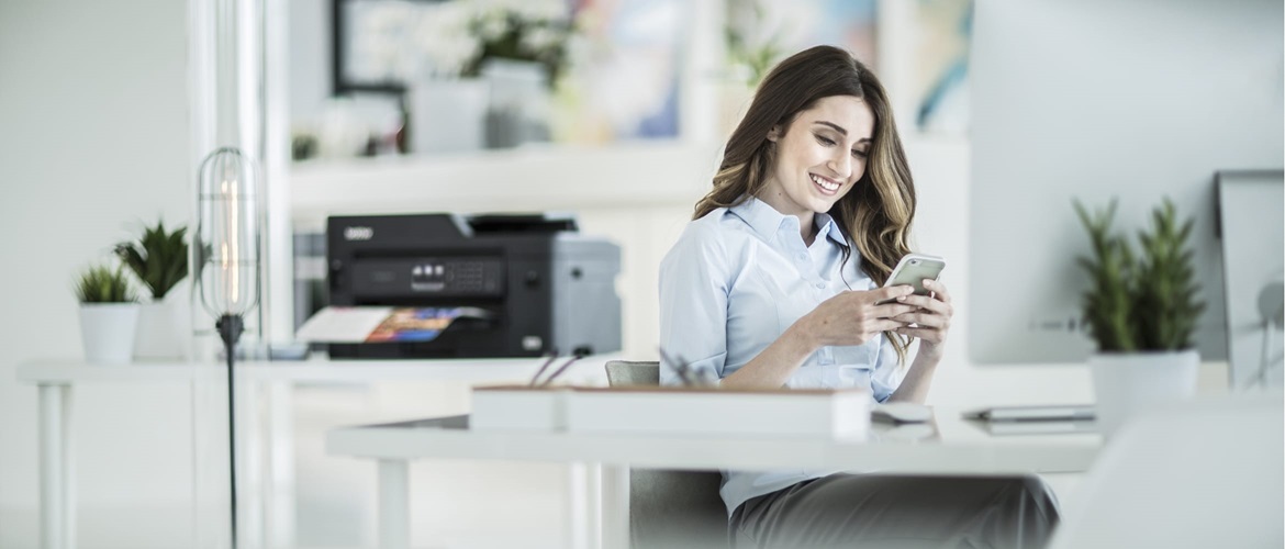Eine Frau sitzt an ihrem Schreibtisch und schaut auf ihr Smartphone. Im Hintergrund ist ein Drucker auf einer Kommode zu sehen.