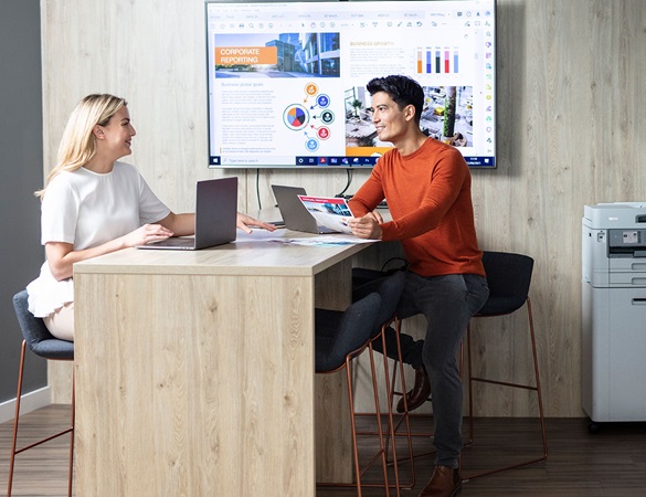 Mann und Frau sitzen an einem Tisch mit Fernseher und Drucker an der Rückwand