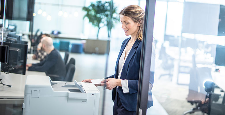 Frau in großem Büro mit Brother-Drucker mit NFC-Karte, PC-Monitoren, Pflanzen, Stühlen
